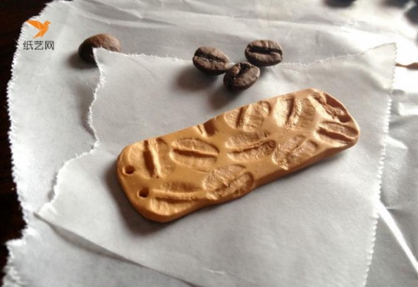 先用超轻粘土做成一个方形的片状之后，就可以用咖啡豆在上面印上花纹了，在两边都打上孔